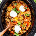 A slow cooker filled with cheesy casserole topped with beans, sour cream, cilantro, and a wooden spoon inside.