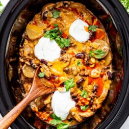 A slow cooker filled with cheesy casserole topped with beans, sour cream, cilantro, and a wooden spoon inside.