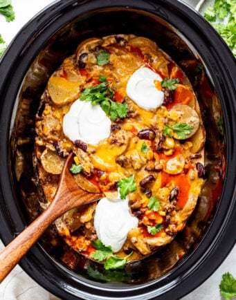 A slow cooker filled with cheesy casserole topped with beans, sour cream, cilantro, and a wooden spoon inside.