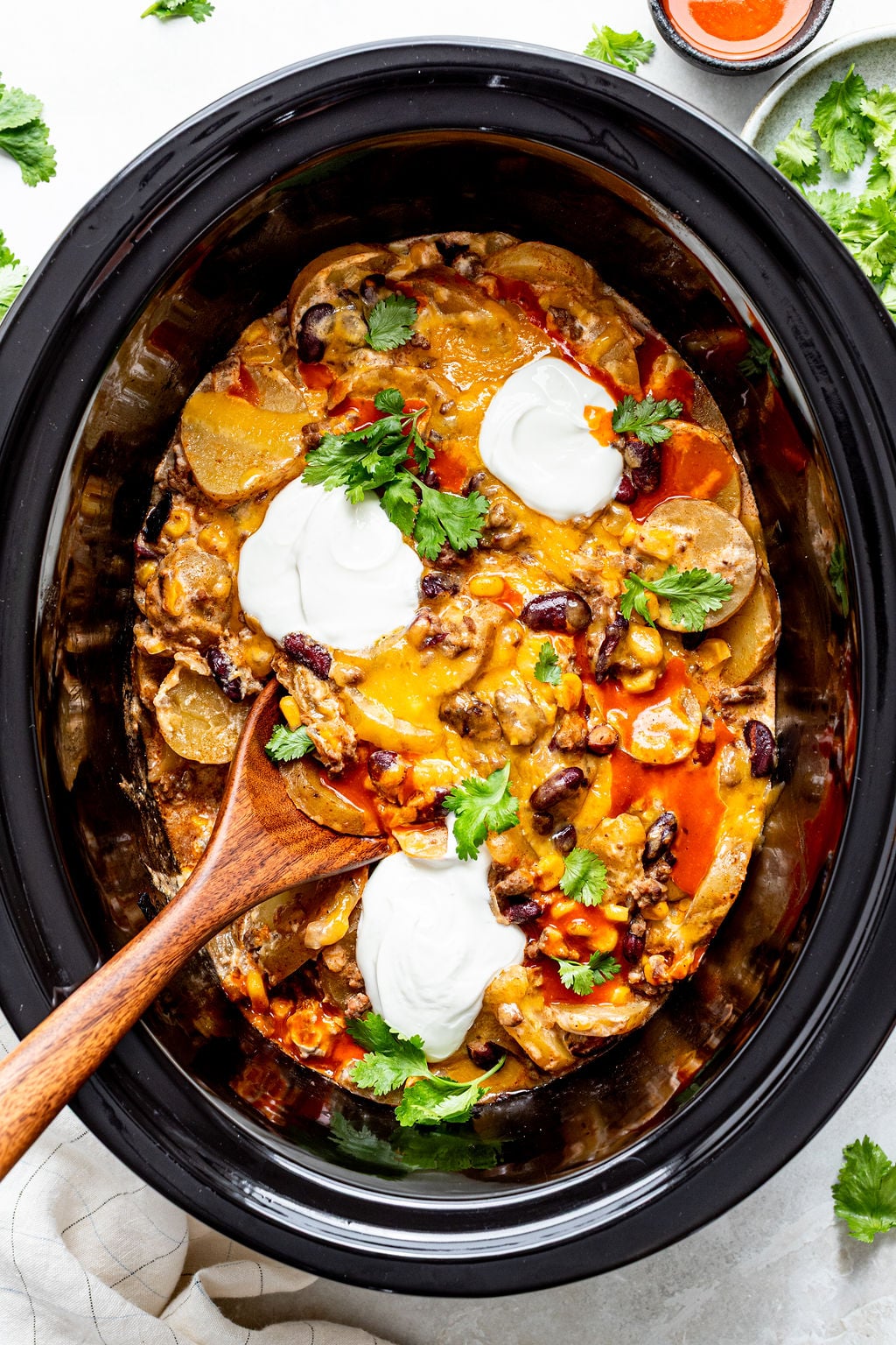 A slow cooker filled with cheesy casserole topped with beans, sour cream, cilantro, and a wooden spoon inside.