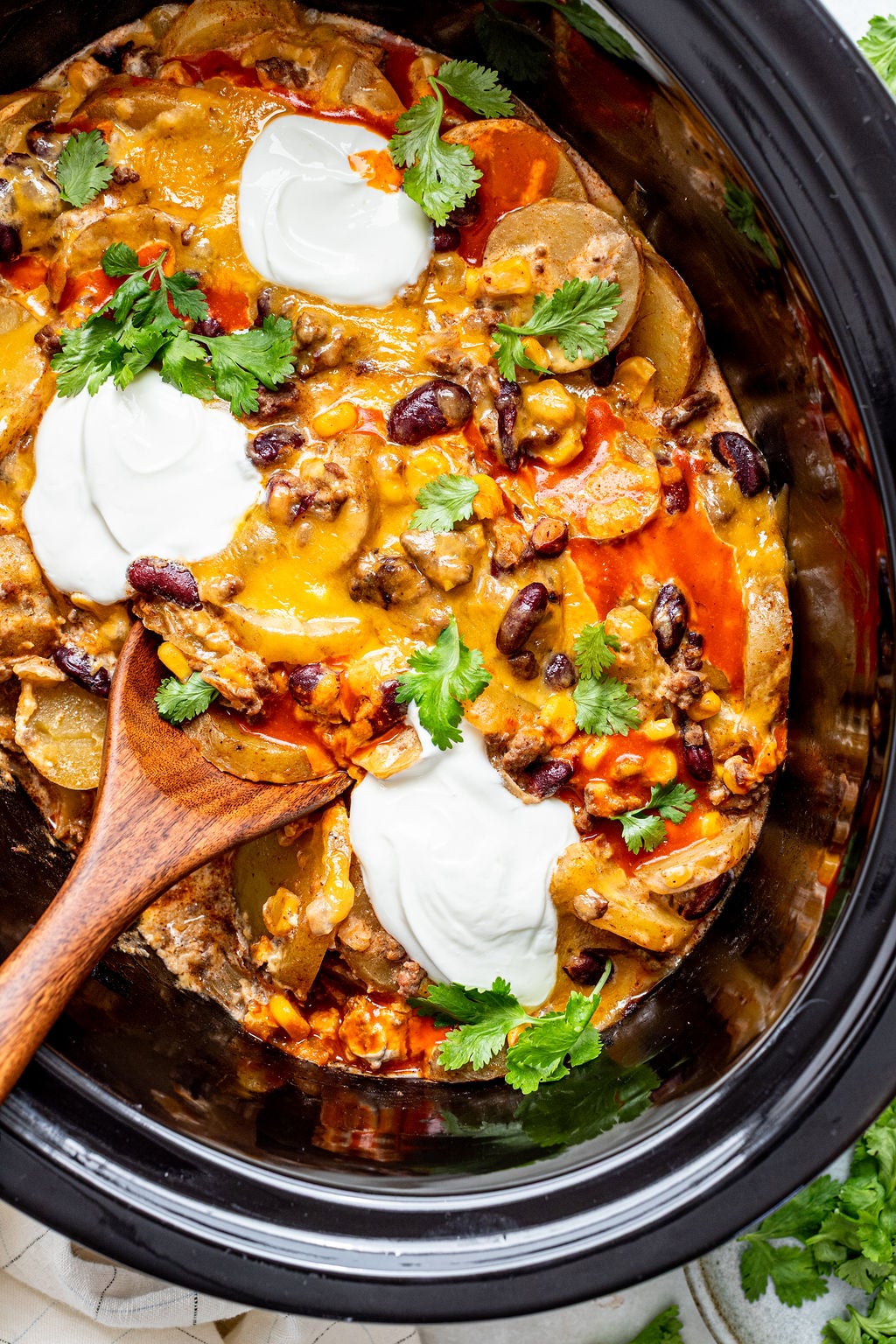 A slow cooker filled with a cheesy casserole topped with sour cream, cilantro, beans, and sliced potatoes, with a wooden spoon resting inside.
