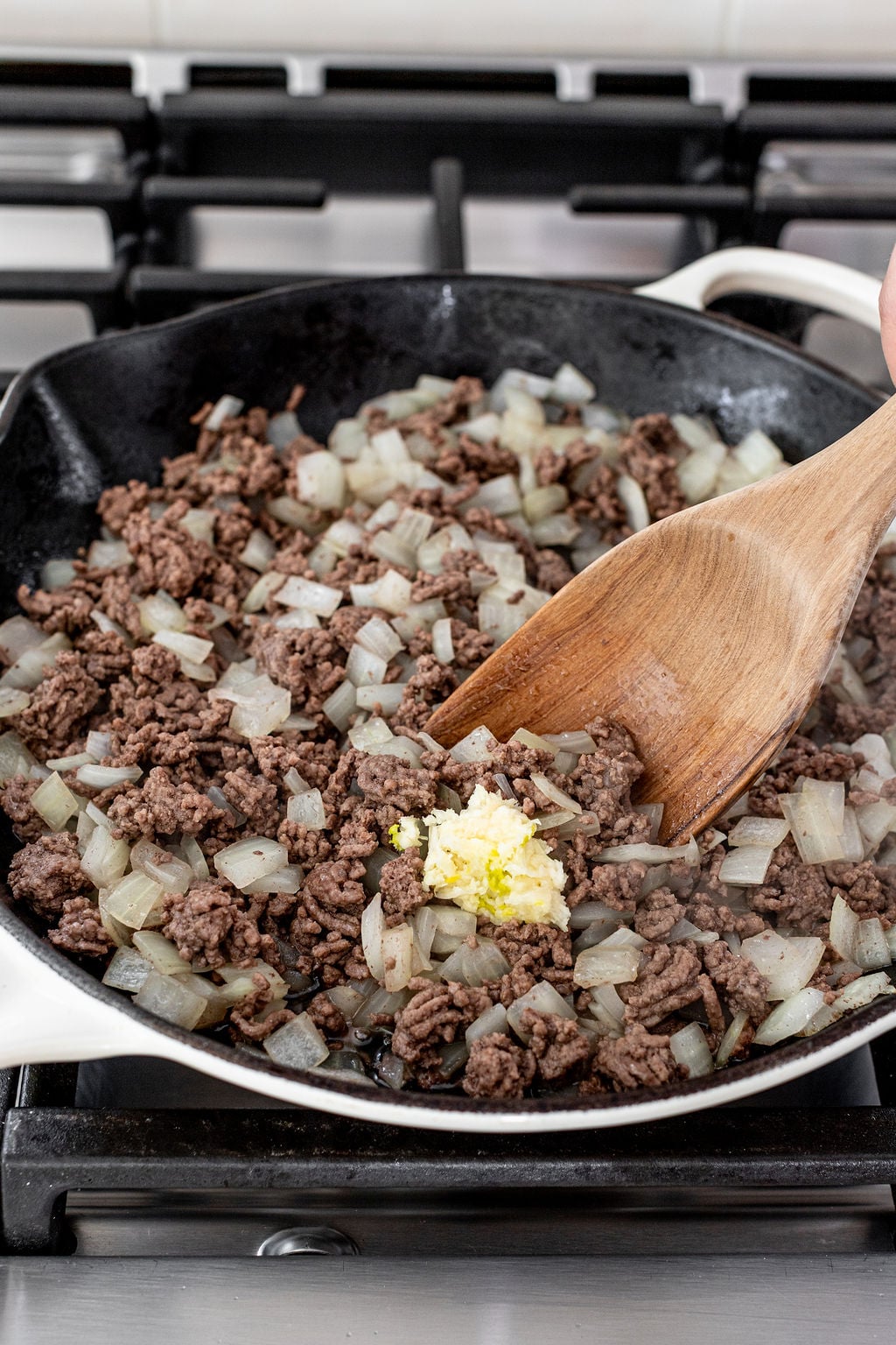 Ground beef and chopped onions are being cooked in a skillet on a stove, with minced garlic added, and a wooden spatula stirring the mixture.