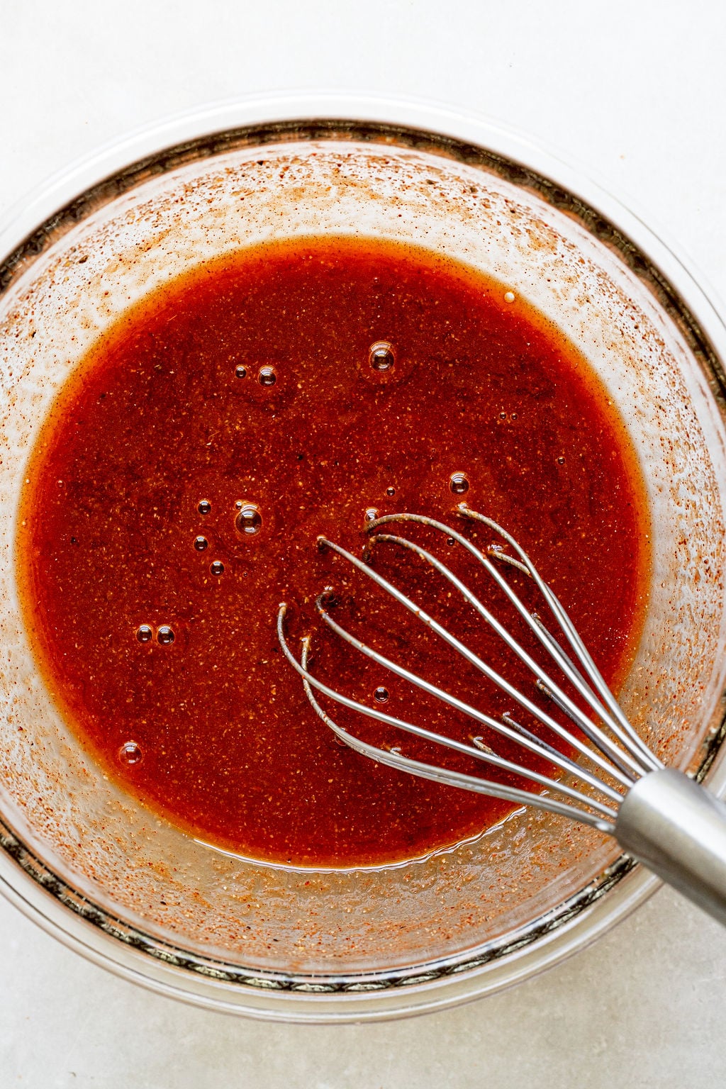A metal whisk rests in a glass bowl filled with a reddish-brown liquid sauce.