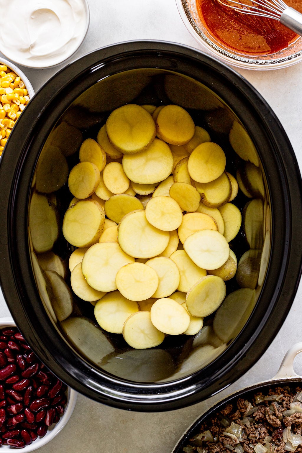 Sliced potatoes are layered in a black slow cooker, surrounded by bowls of corn, kidney beans, sour cream, sauce, and cooked ground meat on a light surface.