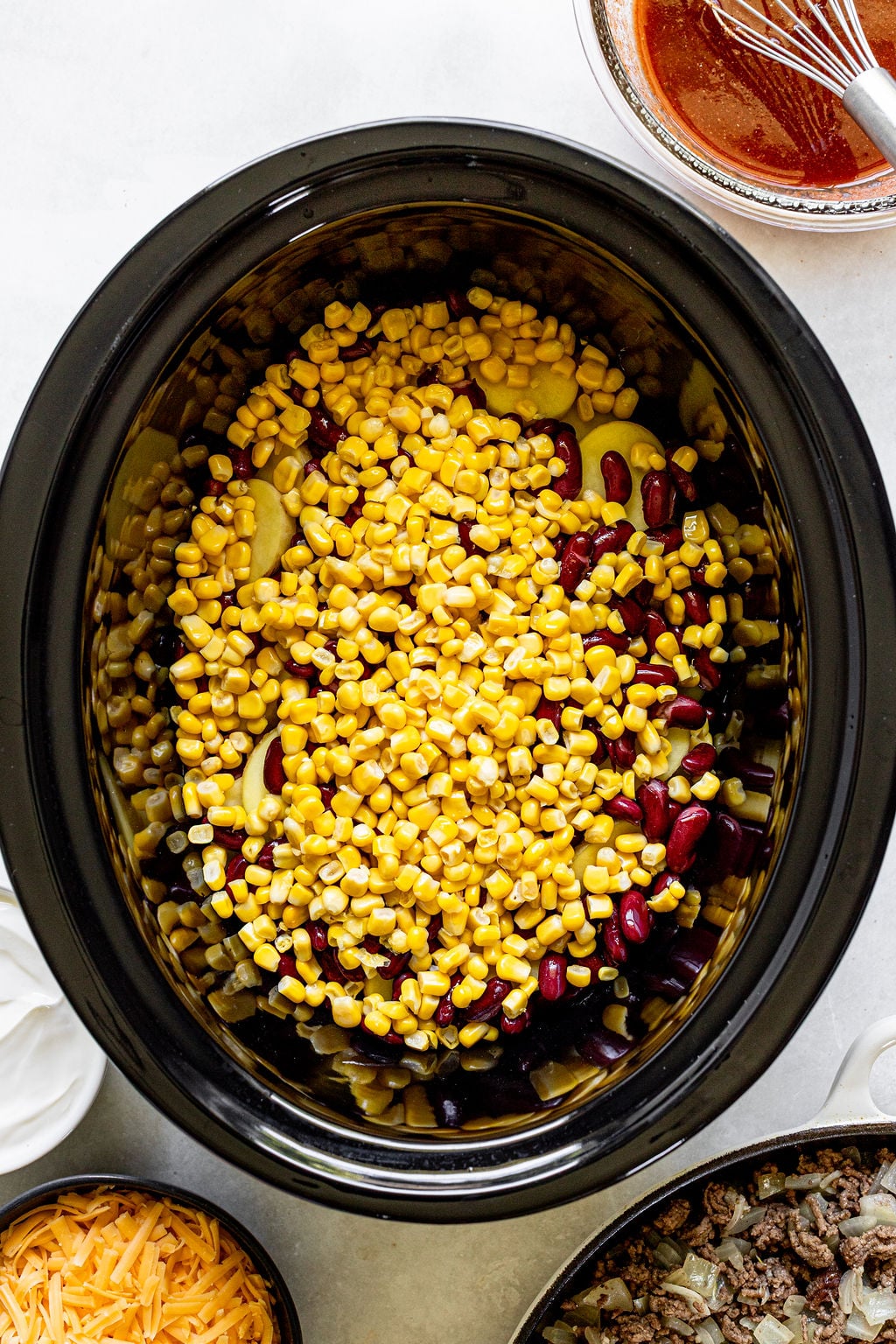 Overhead view of a slow cooker filled with corn, kidney beans, and other ingredients, surrounded by bowls of sauce, cheese, and cooked ground meat.
