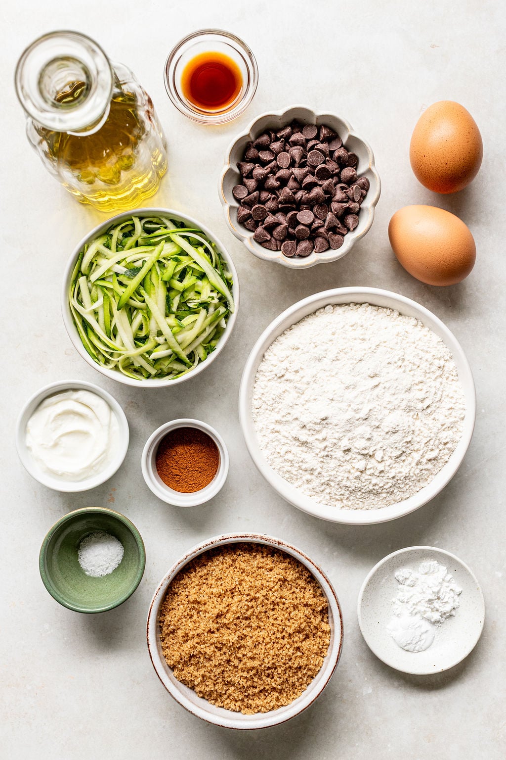 Baking ingredients on a white surface include flour, brown sugar, zucchini, chocolate chips, eggs, oil, vanilla, yogurt, cinnamon, baking powder, and salt in separate bowls.