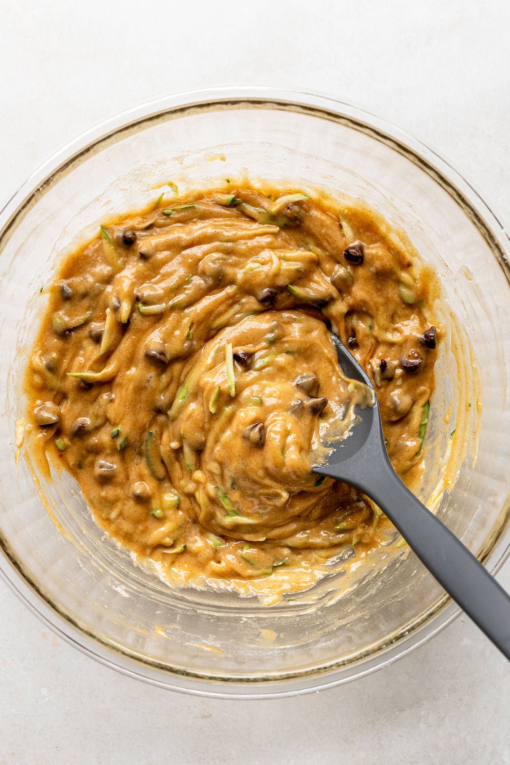 A glass bowl filled with thick, brownish batter mixed with chocolate chips and shredded zucchini, with a black spatula resting inside.