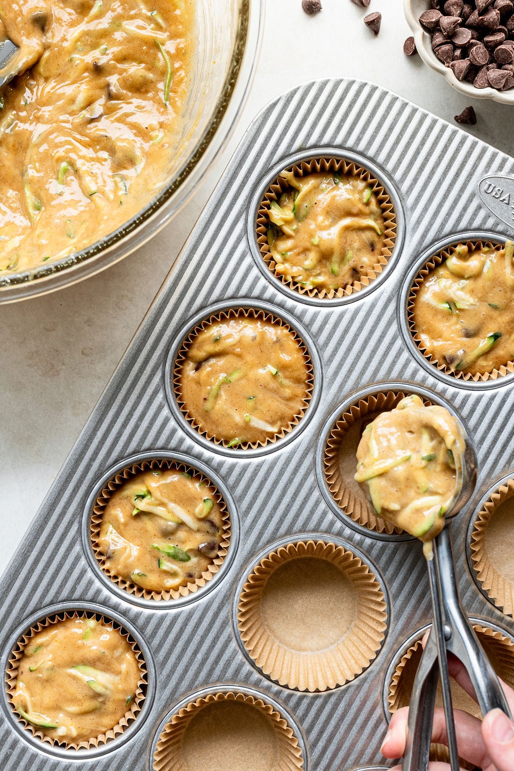 Muffin batter with visible zucchini and chocolate chips being scooped into a lined muffin tin, with a bowl of batter and chocolate chips nearby.