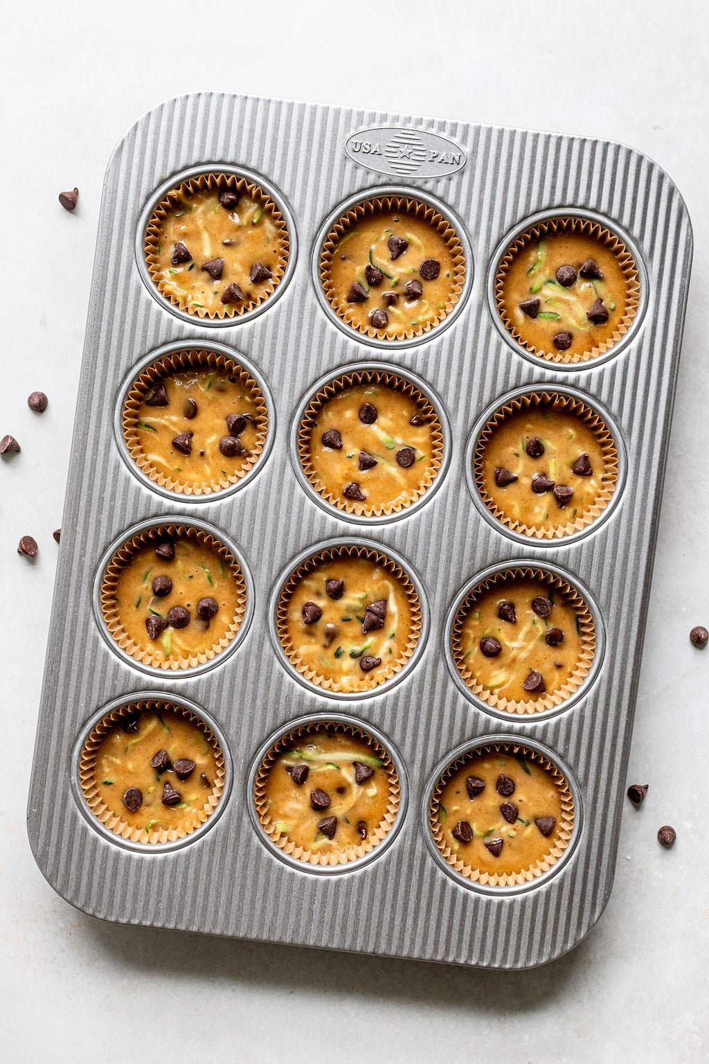 A metal muffin tray with twelve unbaked zucchini chocolate chip muffins in paper liners, sprinkled with chocolate chips, on a light surface.