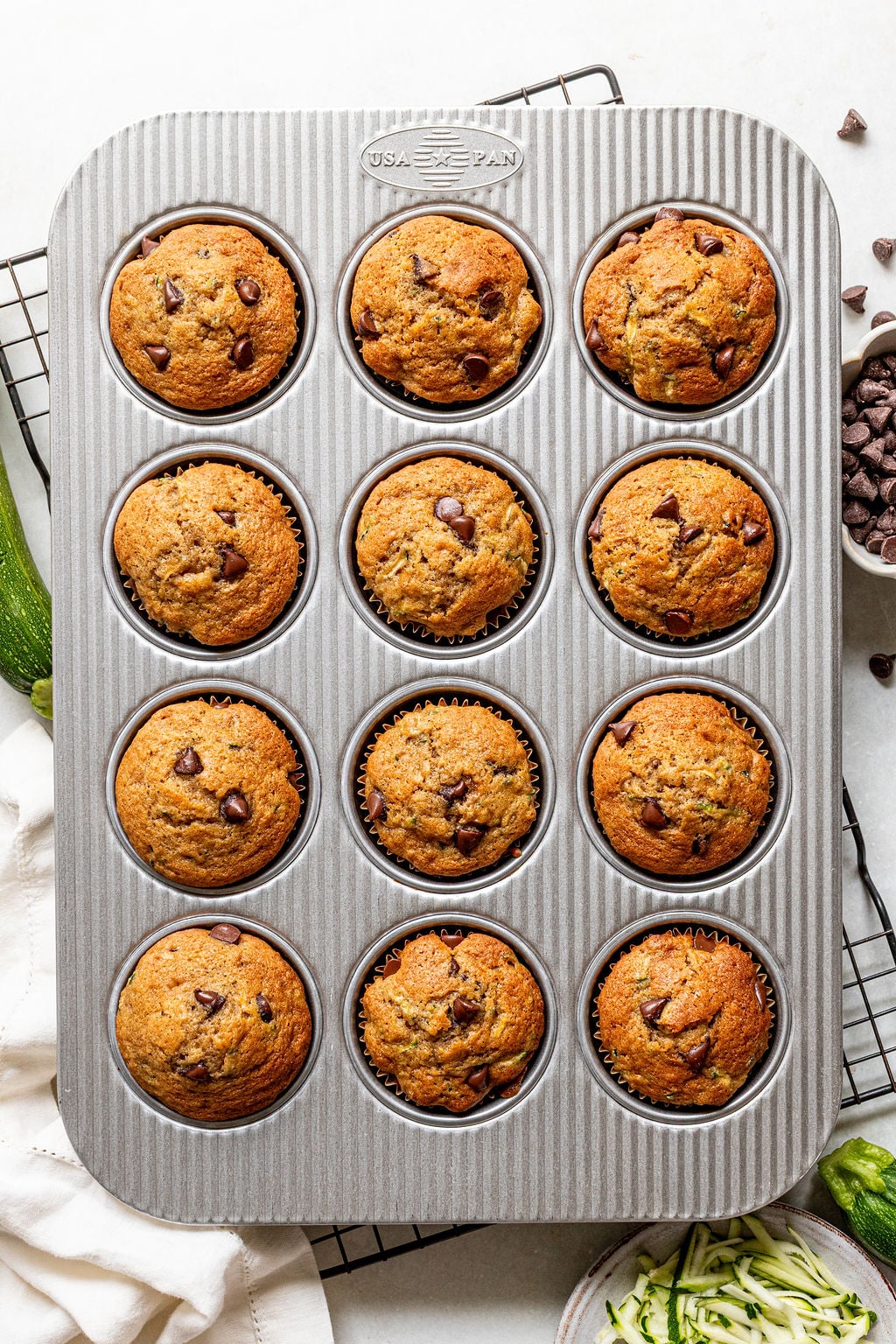 A metal muffin tin filled with twelve baked chocolate chip muffins sits on a cooling rack, with a bowl of chocolate chips and some shredded zucchini nearby.