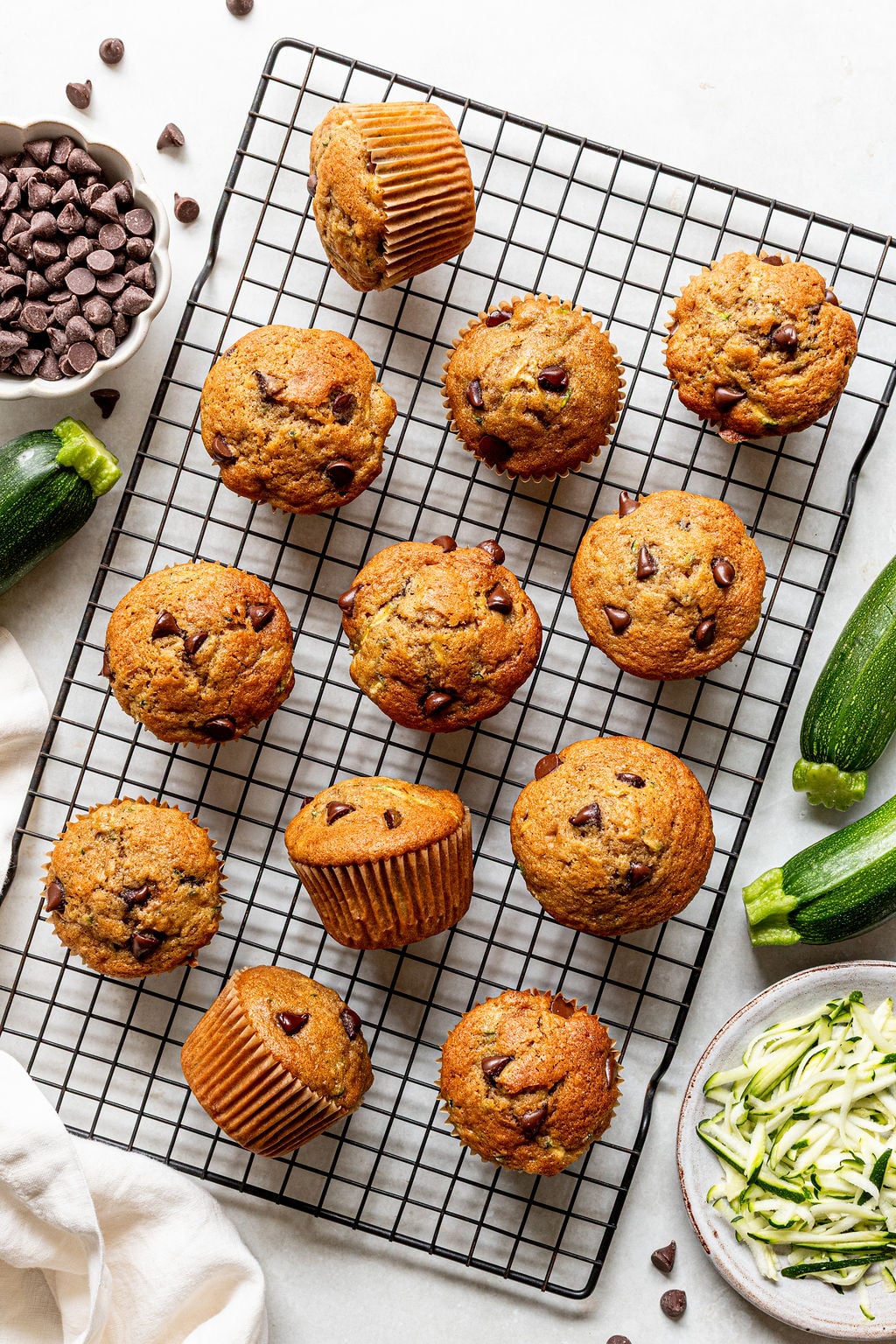 Twelve chocolate chip muffins are cooling on a wire rack, surrounded by chocolate chips, whole zucchinis, a bowl of shredded zucchini, and a white napkin on a light surface.