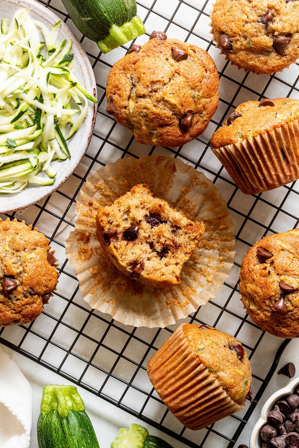 Chocolate chip zucchini muffins are arranged on a cooling rack, with one muffin cut in half. A bowl of shredded zucchini and whole zucchinis are also visible.