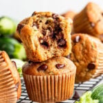 A stack of chocolate chip zucchini muffins sits on a cooling rack, with the top muffin broken open to show the chocolate chips inside.