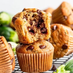 A stack of chocolate chip zucchini muffins sits on a cooling rack, with the top muffin broken open to show the chocolate chips inside.