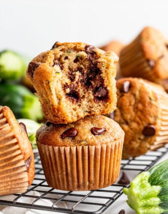 A stack of chocolate chip zucchini muffins sits on a cooling rack, with the top muffin broken open to show the chocolate chips inside.