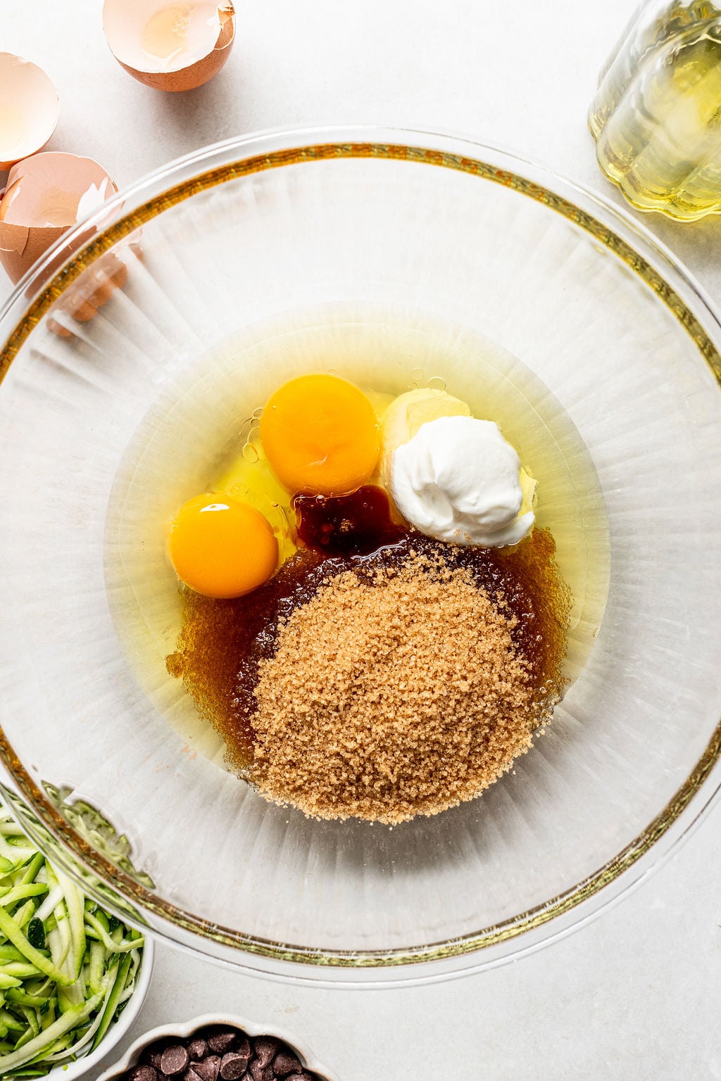 A glass bowl contains eggs, brown sugar, oil, vanilla, and sour cream. Eggshells, shredded zucchini, chocolate chips, and oil are arranged around the bowl.