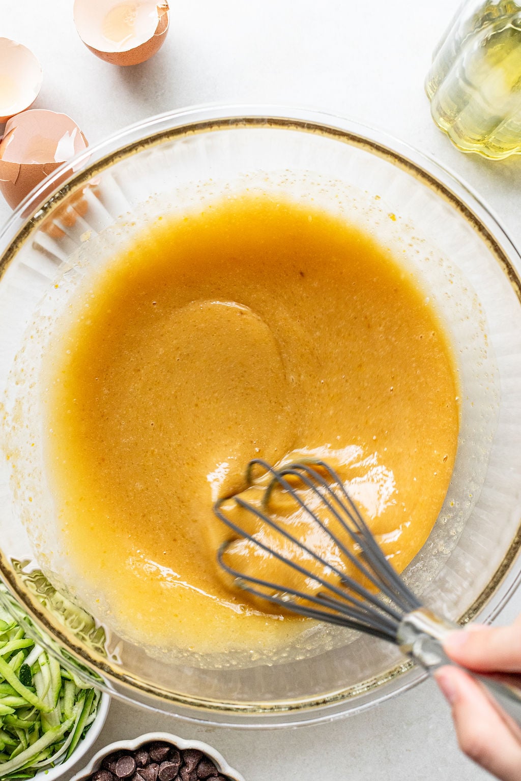 A hand uses a whisk to mix a yellow batter in a glass bowl, surrounded by eggshells, oil, shredded zucchini, and chocolate chips on a white surface.