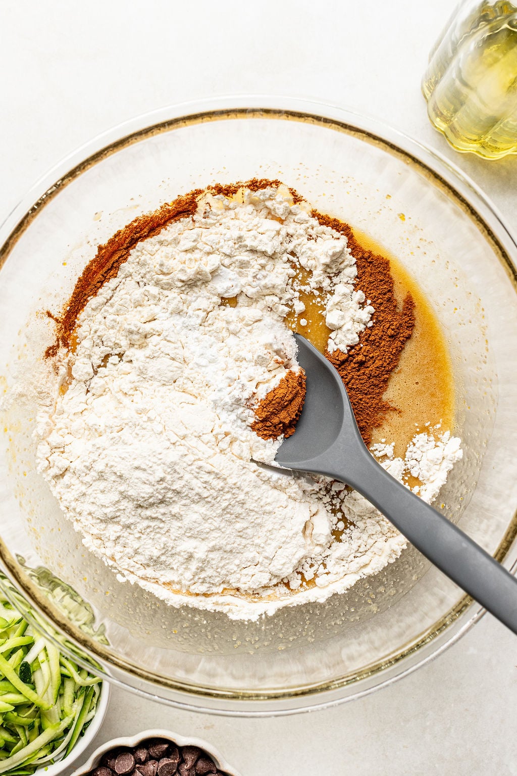 A glass bowl with flour, cinnamon, and wet ingredients being mixed with a gray spoon, surrounded by oil, shredded zucchini, and chocolate chips.
