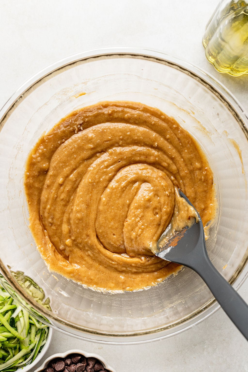 A glass bowl containing brown batter being stirred with a gray spatula; shredded zucchini, chocolate chips, and oil are nearby.