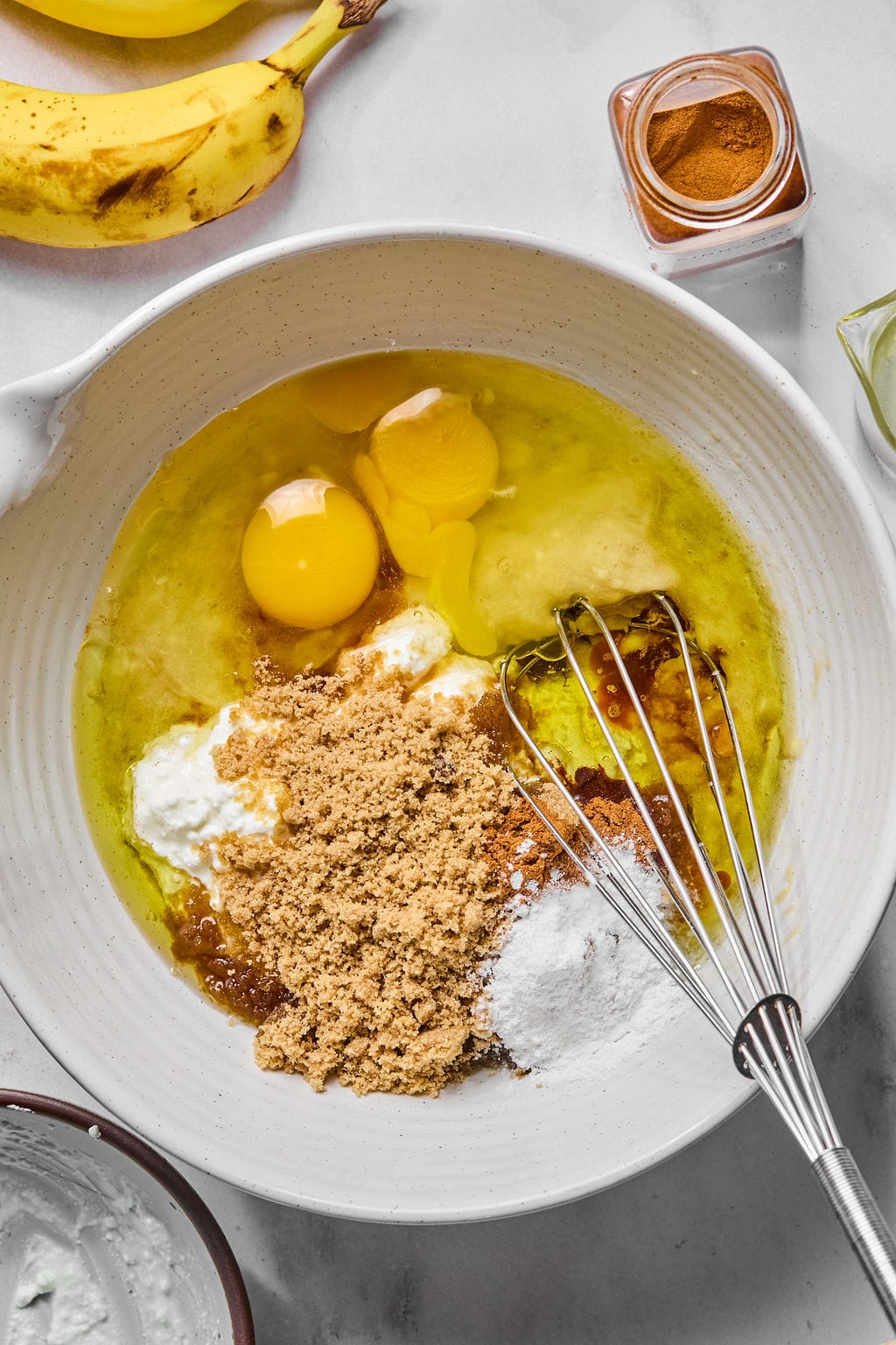 A mixing bowl with eggs, brown sugar, yogurt, and other ingredients, with a whisk beside them. Bananas and a jar of cinnamon are nearby.