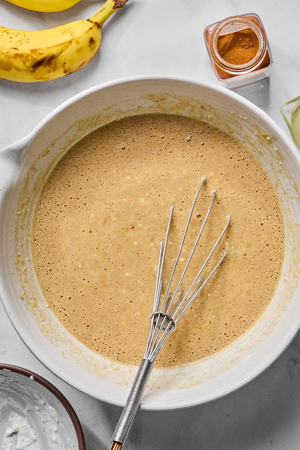 A mixing bowl with banana bread batter and a whisk, surrounded by bananas, a jar of cinnamon, and other baking ingredients on a countertop.