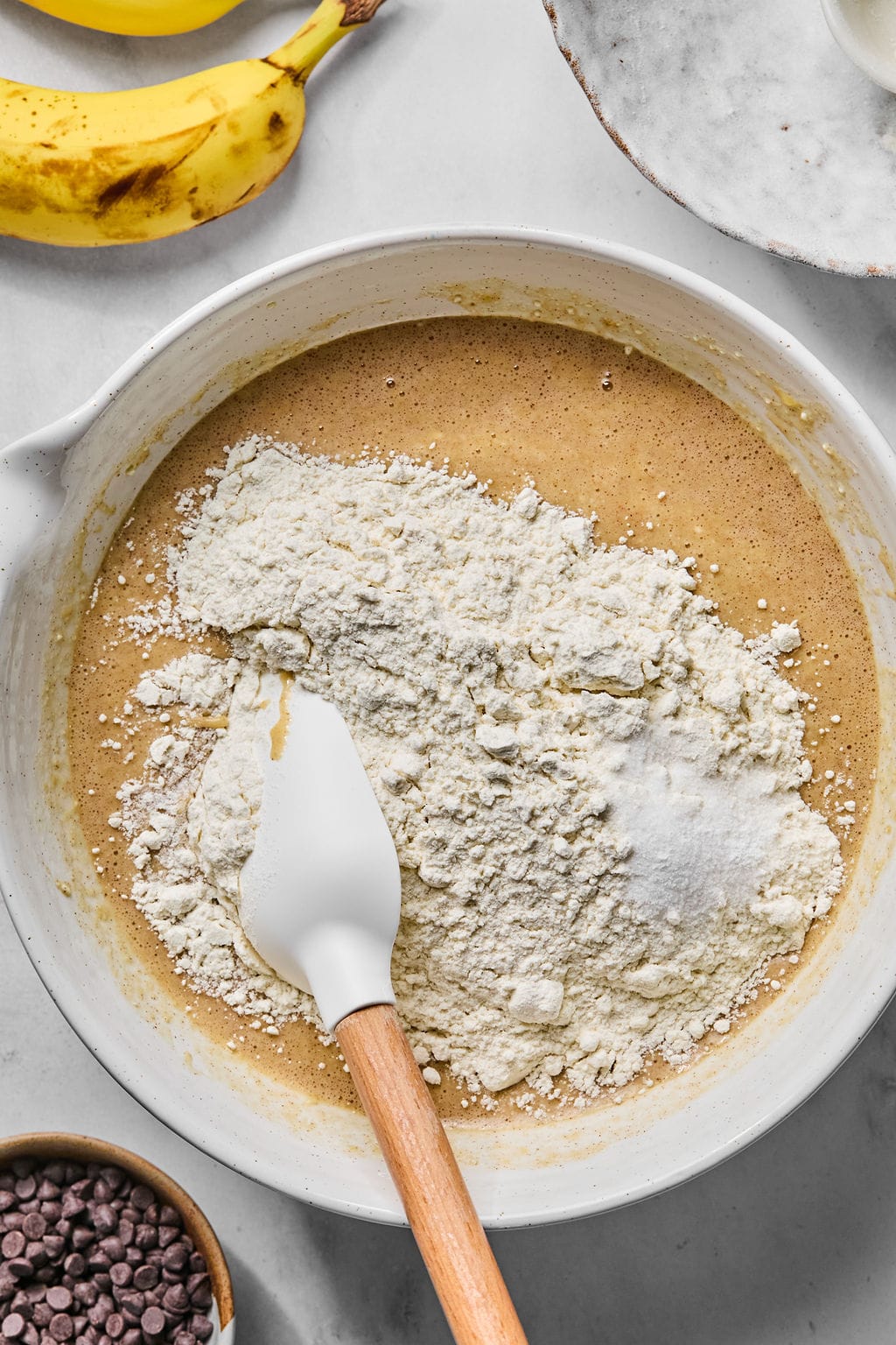 A mixing bowl with flour on top of wet ingredients, a spatula resting inside, and bananas and chocolate chips nearby on the counter.