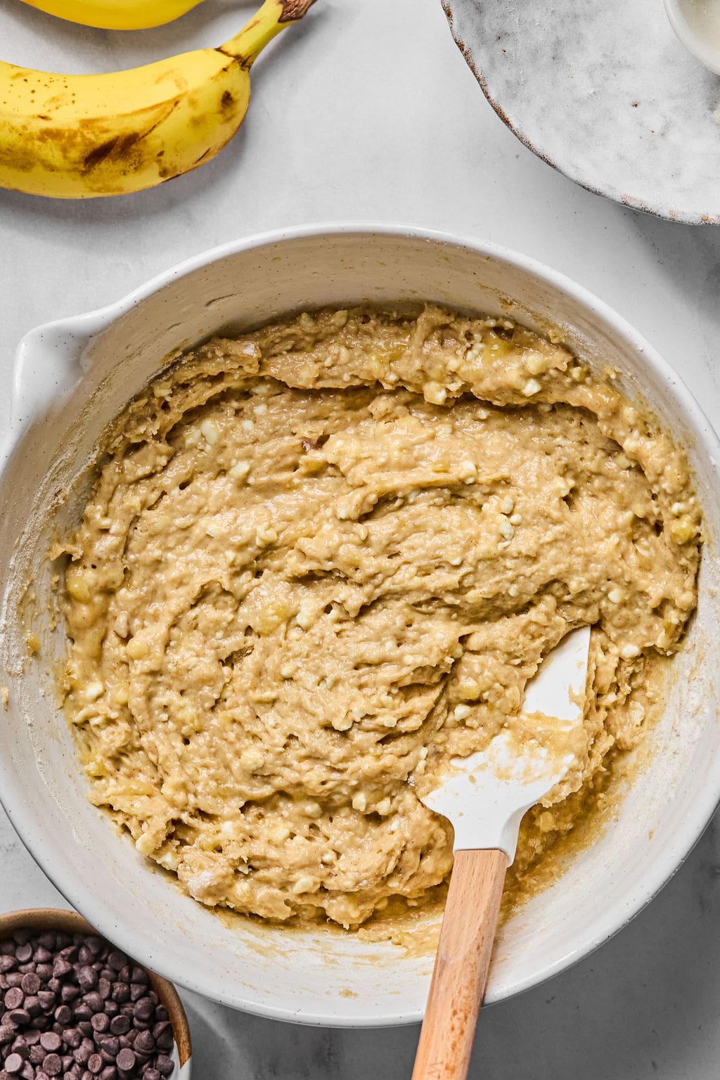 Mixing bowl with banana bread batter and a spatula, surrounded by bananas, a bowl of chocolate chips, and a white plate.