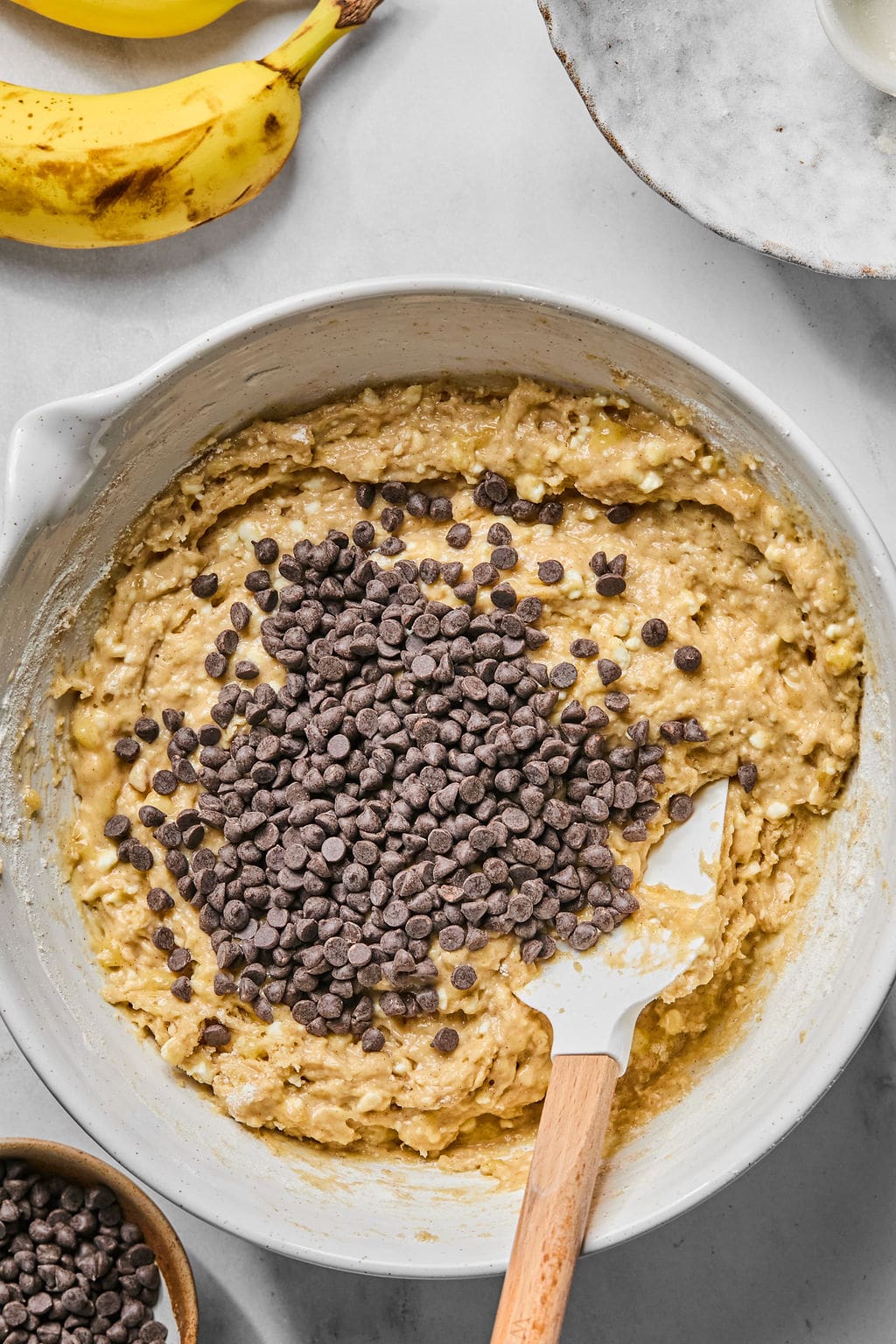 Mixing bowl with banana bread batter and chocolate chips, a spatula is inside, and bananas are visible in the background on a white surface.