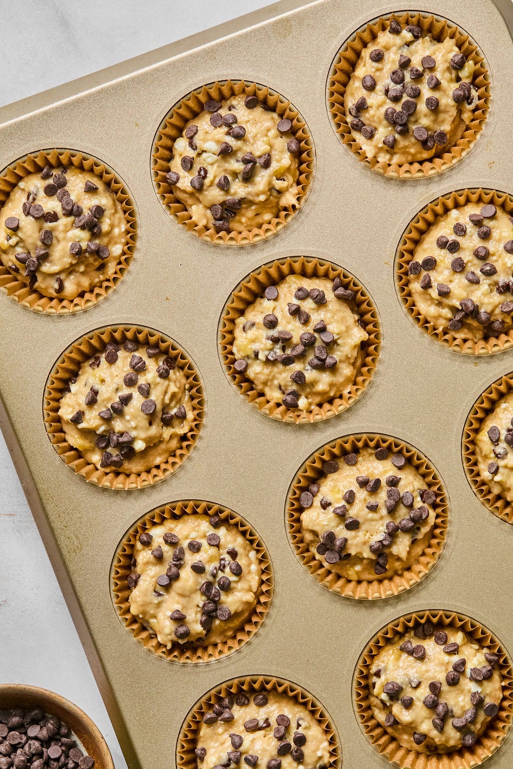 A muffin tin filled with unbaked muffin batter in paper liners, topped with chocolate chips, ready for baking.