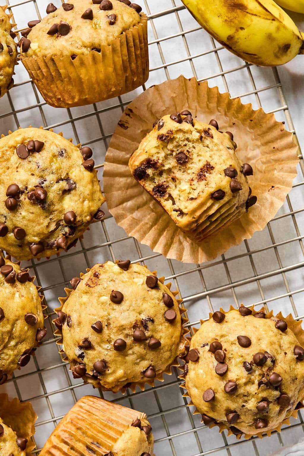 Several banana chocolate chip muffins on a cooling rack, with one muffin partially unwrapped and a bunch of bananas in the corner.