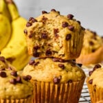 Two chocolate chip banana muffins are stacked on a cooling rack, with one muffin missing a bite. Bananas and more muffins are visible in the background.