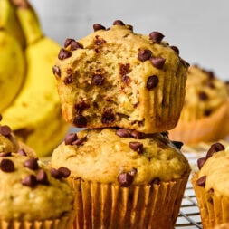 Two chocolate chip banana muffins are stacked on a cooling rack, with one muffin missing a bite. Bananas and more muffins are visible in the background.
