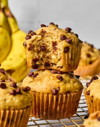 Two chocolate chip banana muffins are stacked on a cooling rack, with one muffin missing a bite. Bananas and more muffins are visible in the background.