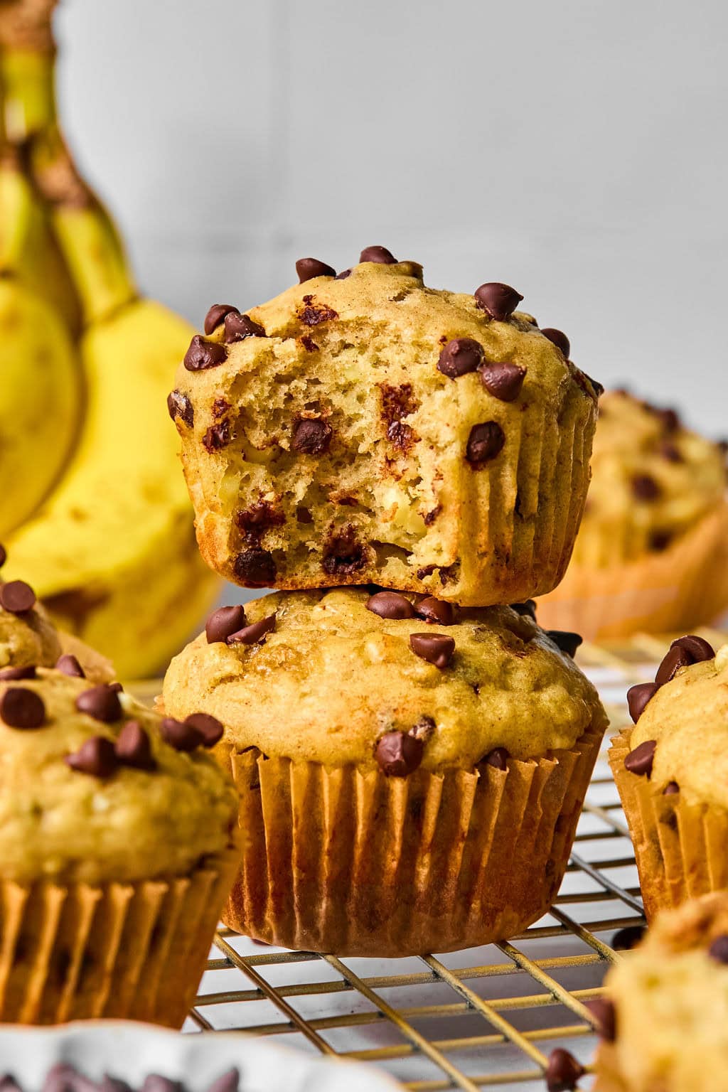 Two chocolate chip banana muffins are stacked on a cooling rack, with one muffin missing a bite. Bananas and more muffins are visible in the background.