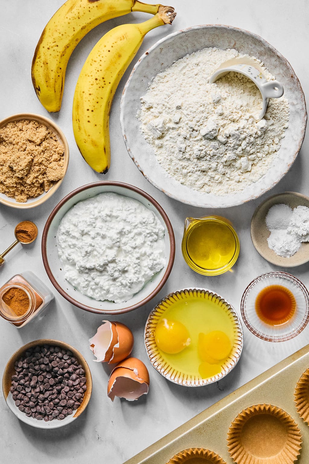 A flat lay of ingredients for baking, including bananas, flour, eggs, brown sugar, cottage cheese, oil, chocolate chips, vanilla, baking powder, cinnamon, and cupcake liners.