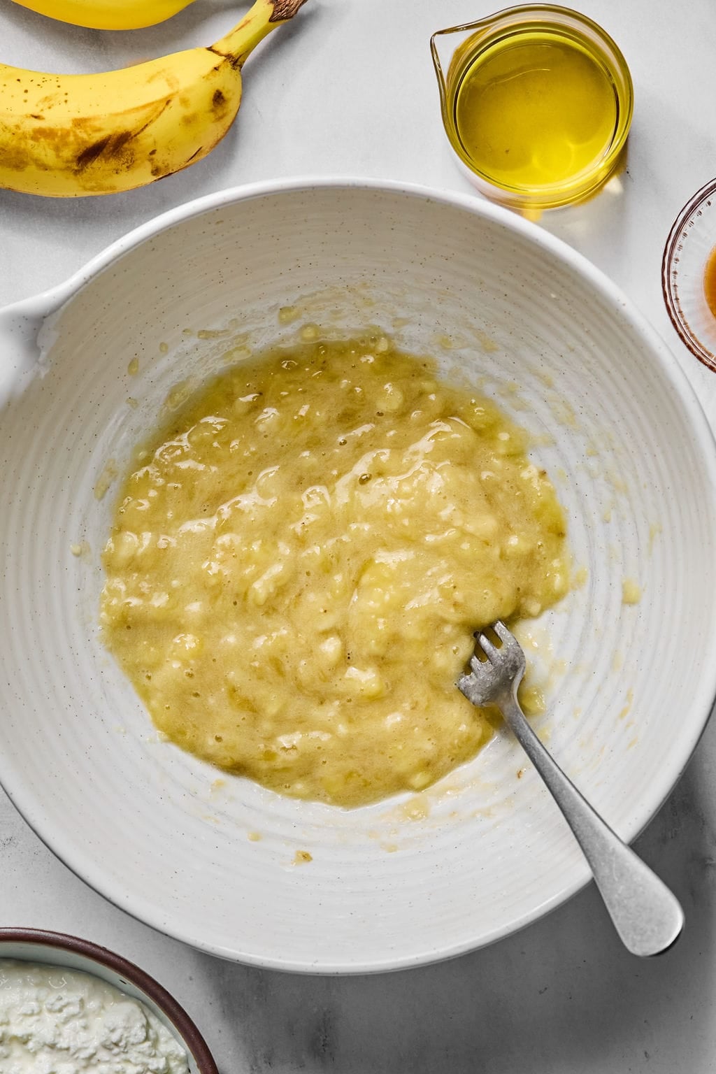 A white bowl with mashed bananas and a fork. Surrounding the bowl are a banana, a small glass pitcher of oil, and partial views of other ingredients on a white surface.