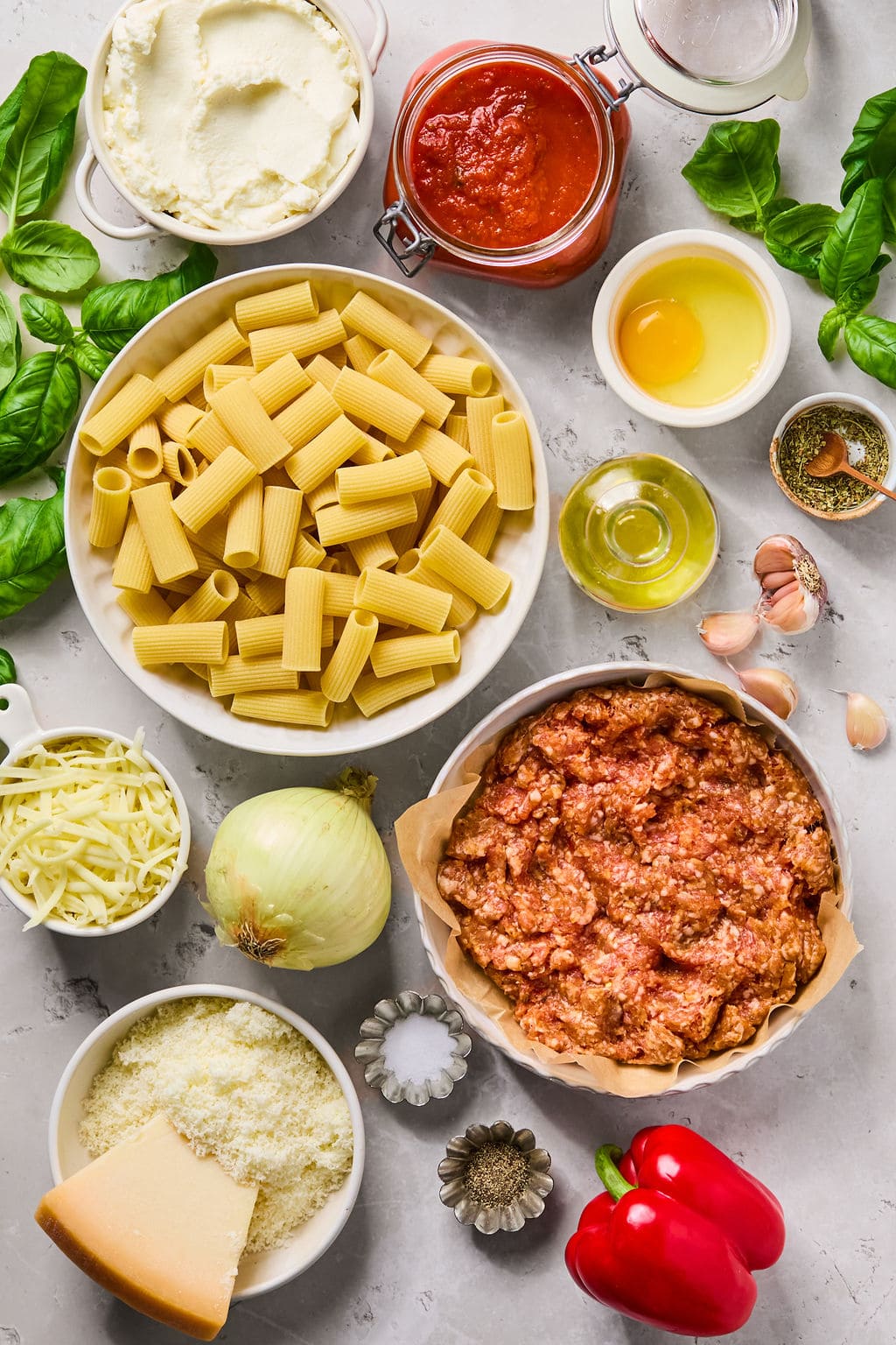 Various ingredients including rigatoni, ricotta, mozzarella, Parmesan, ground meat, tomato sauce, eggs, onion, pepper, basil leaves, garlic cloves, oil, and seasonings on a table.