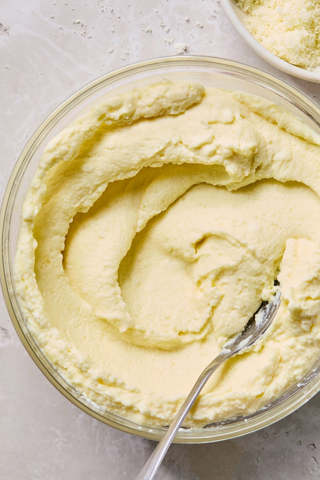 A glass bowl filled with smooth, whipped ricotta cheese sits on a light surface with a spoon resting inside.