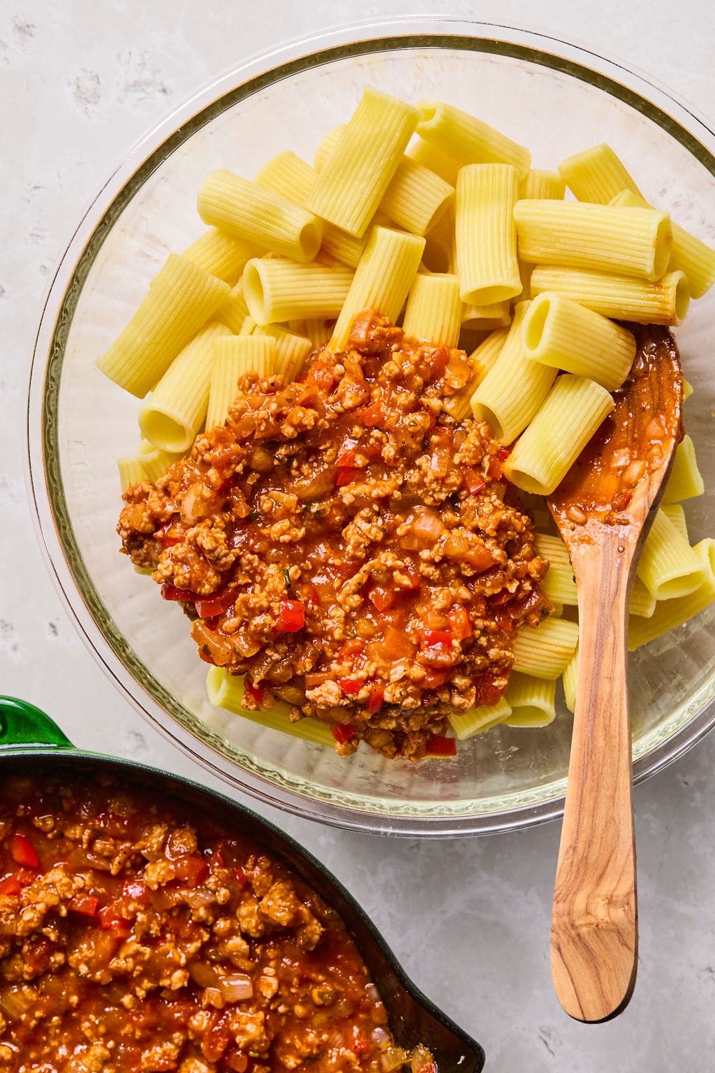 Cooked rigatoni pasta in a glass bowl is topped with meat sauce. A wooden spoon rests on the bowl, and a pan with more sauce is partially visible nearby.