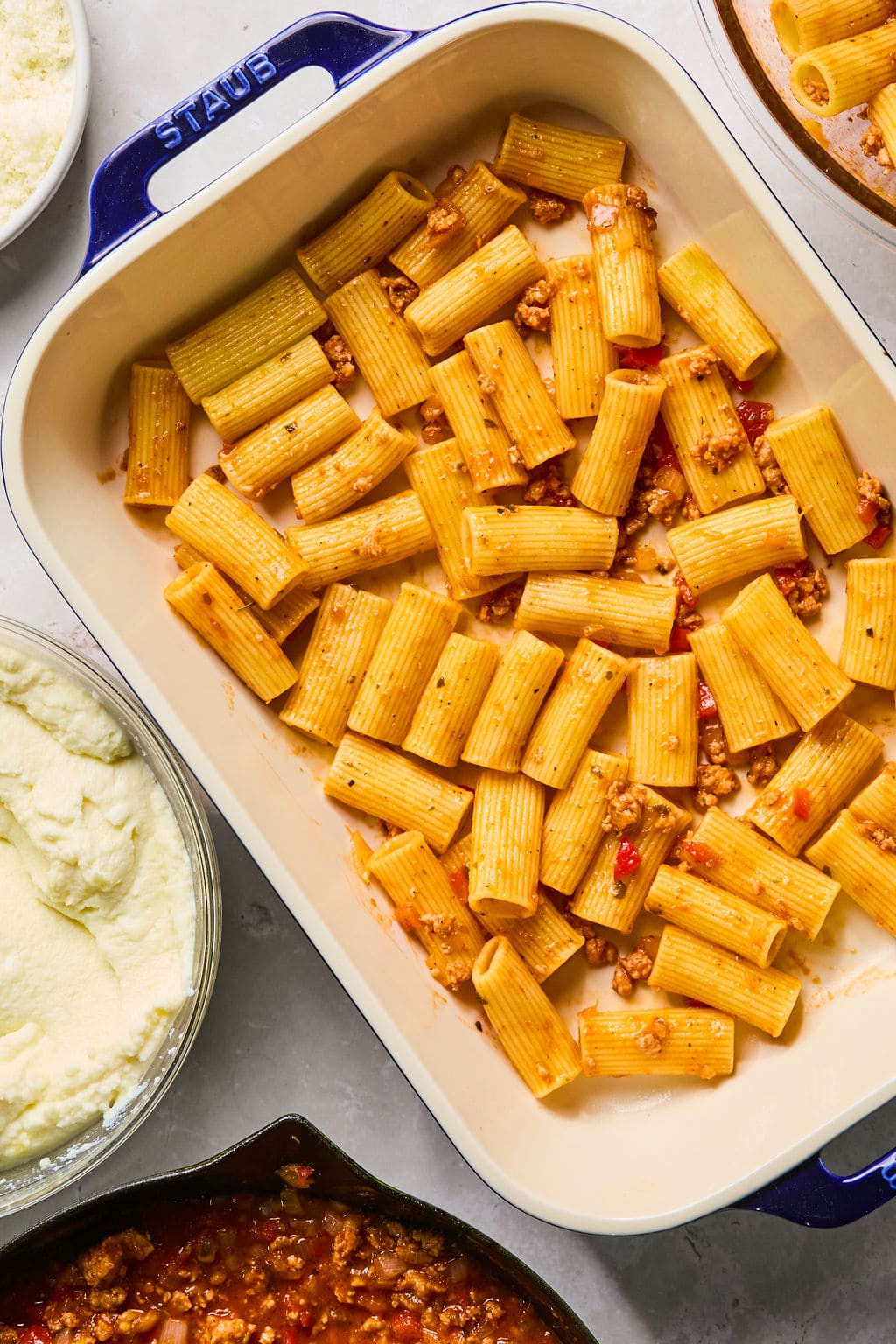 Baking dish with rigatoni pasta and meat sauce, surrounded by bowls of ricotta cheese and additional meat sauce on a countertop.