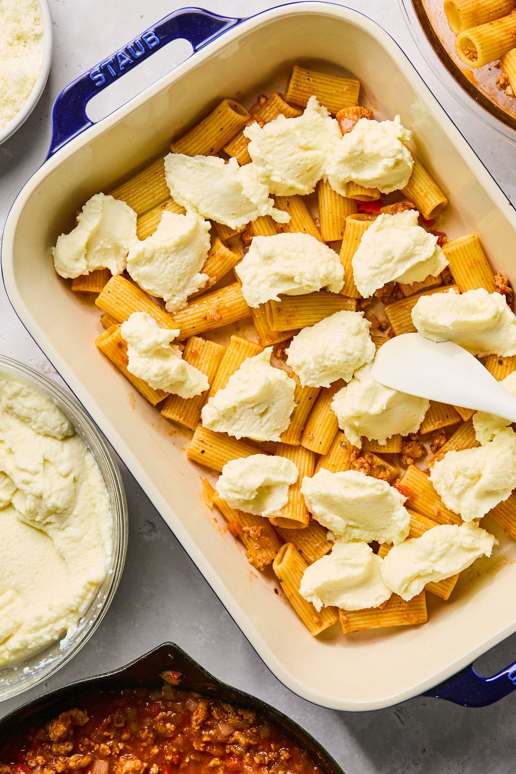 Rectangular baking dish with rigatoni pasta, meat sauce, and dollops of ricotta cheese being spread with a spatula; ingredients and sauces are arranged around the dish.