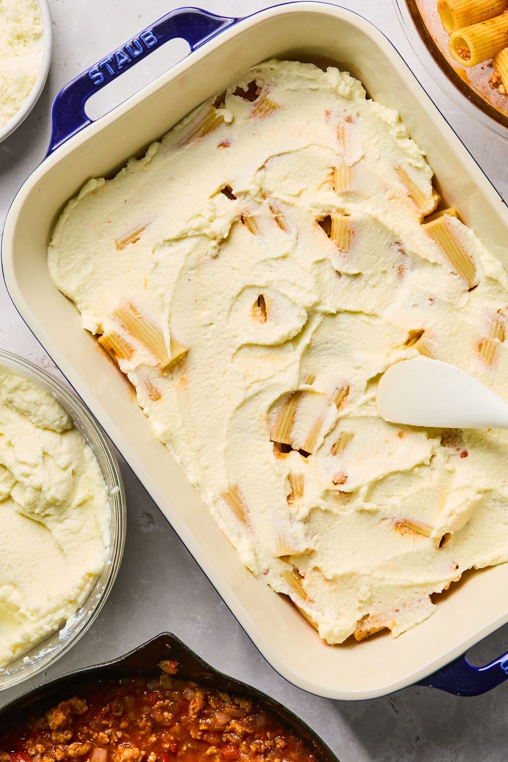A baking dish filled with rigatoni pasta topped with a layer of creamy ricotta cheese mixture, being spread with a spatula.