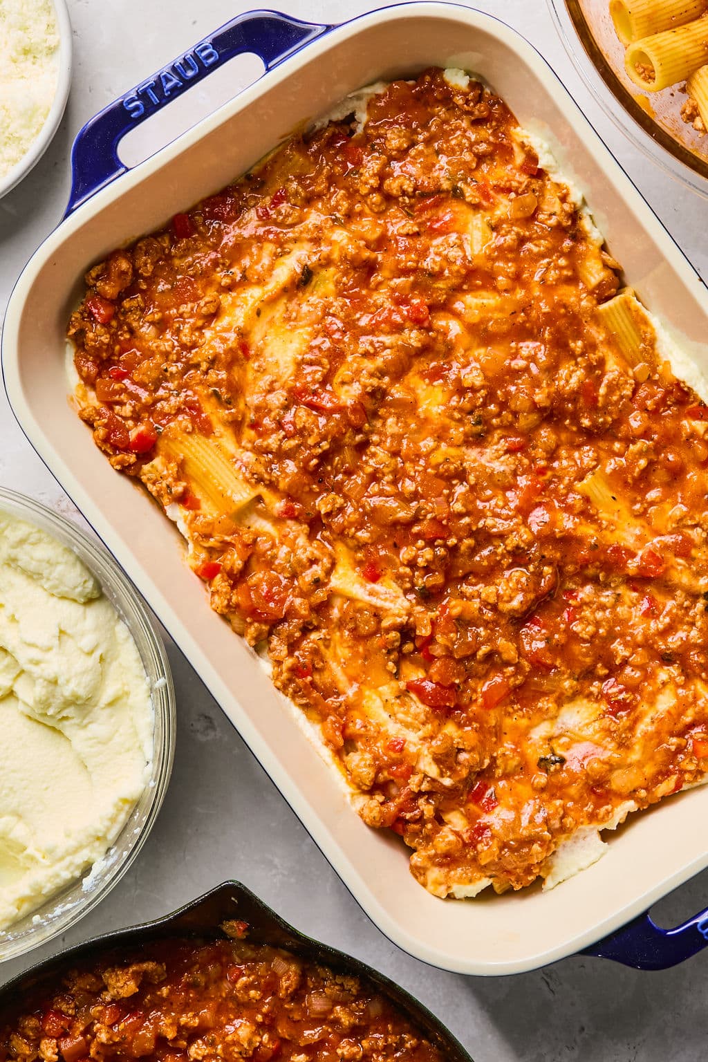 A baking dish filled with lasagna featuring layers of pasta, meat sauce, and cheese, shown from above. Surrounding the dish are bowls of ricotta, cooked pasta, and extra sauce.
