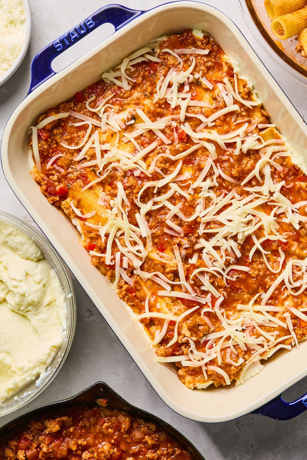 Rectangular casserole dish filled with layers of pasta, meat sauce, ricotta cheese, and shredded mozzarella, ready to be baked. Surrounding the dish are bowls with ingredients.