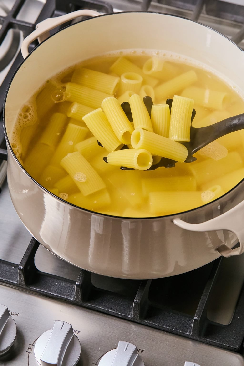 A black ladle lifts rigatoni pasta from a pot of boiling water on a stovetop.