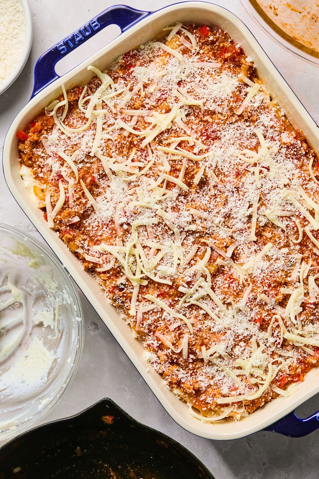 A baking dish filled with assembled pasta bake topped with shredded cheese and sauce, ready to be baked. Surrounding are bowls with ingredients and utensils.