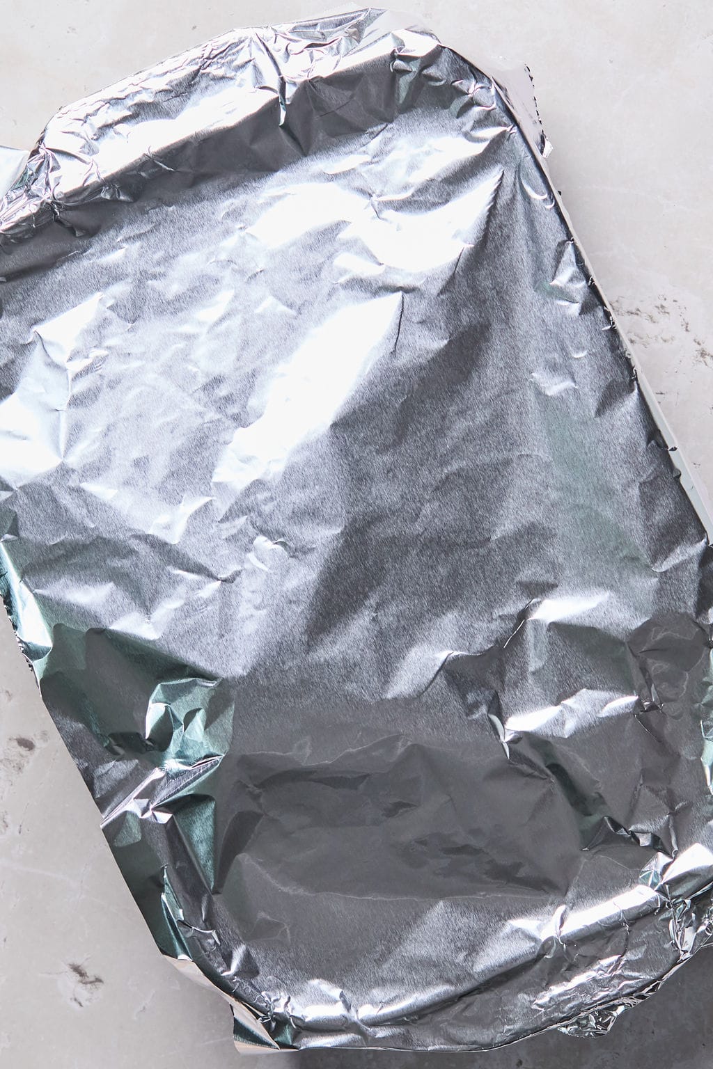 A rectangular baking dish covered with crinkled aluminum foil is placed on a light gray surface.