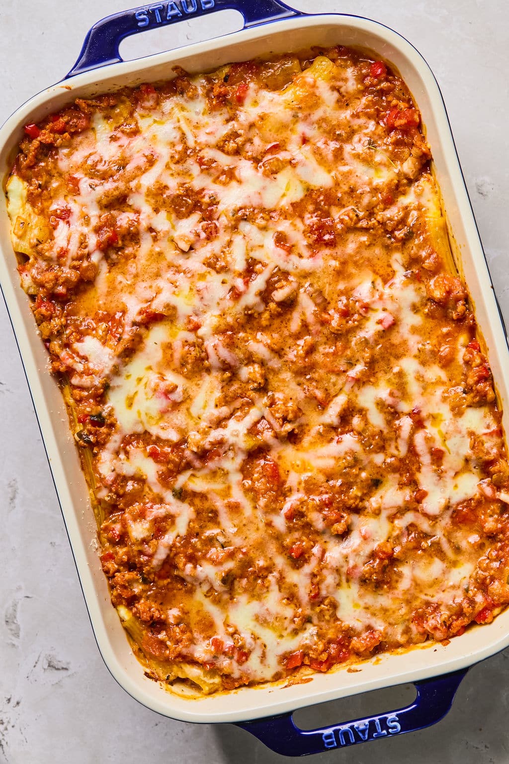 Baked paste in a rectangular ceramic dish with visible layers of melted cheese, meat sauce, and pasta, shown on a light-colored surface.