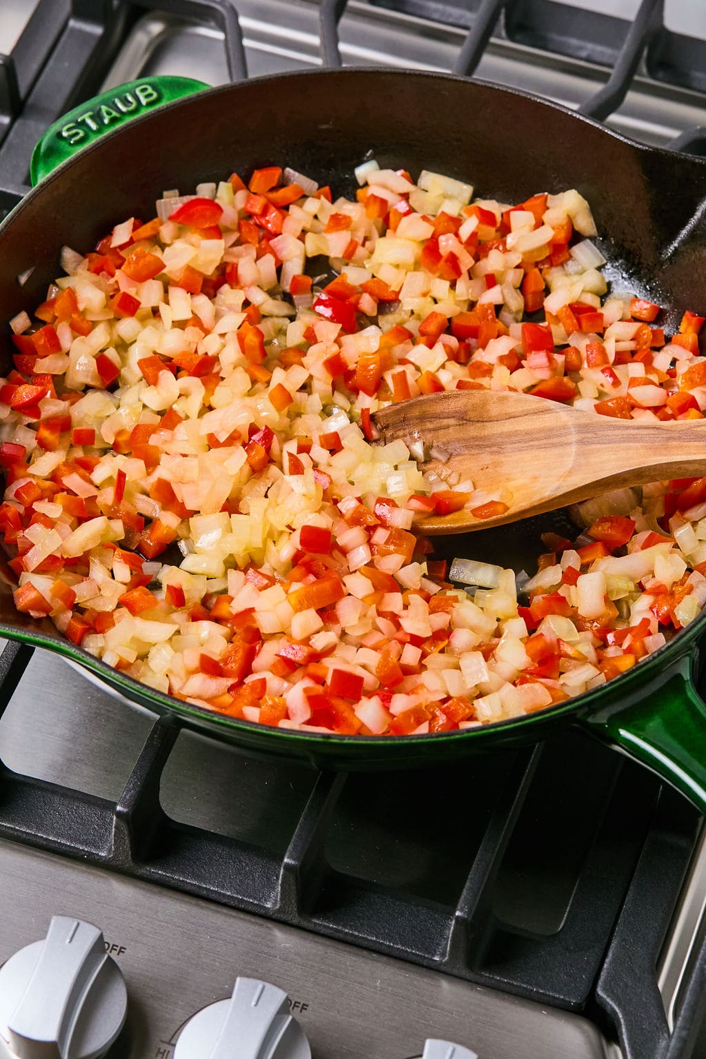 Diced onions and red bell peppers are sautéing in a green skillet on a gas stove, being stirred with a wooden spoon.