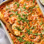 Baked pasta casserole topped with melted cheese and chopped fresh basil, with a wooden spoon serving a portion from a rectangular dish.